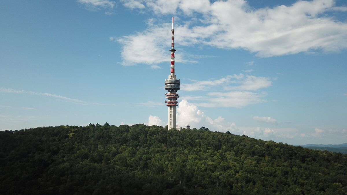 The red and white TV tower of Pécs rising above the forested Misina hill under a blue sky with clouds