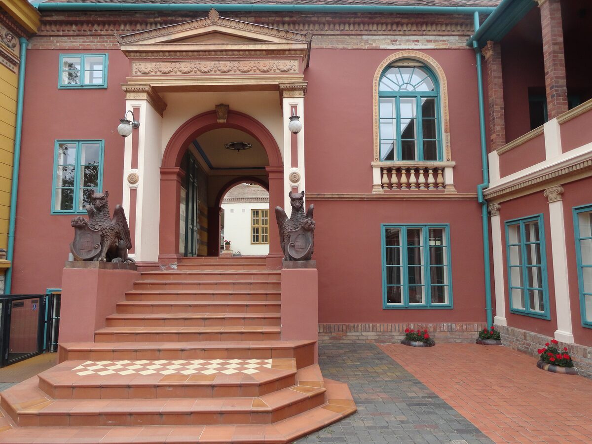 A pink building in the Zsolnay Cultural Quarter with a portico, steps and two ceramic griffins at the entrance