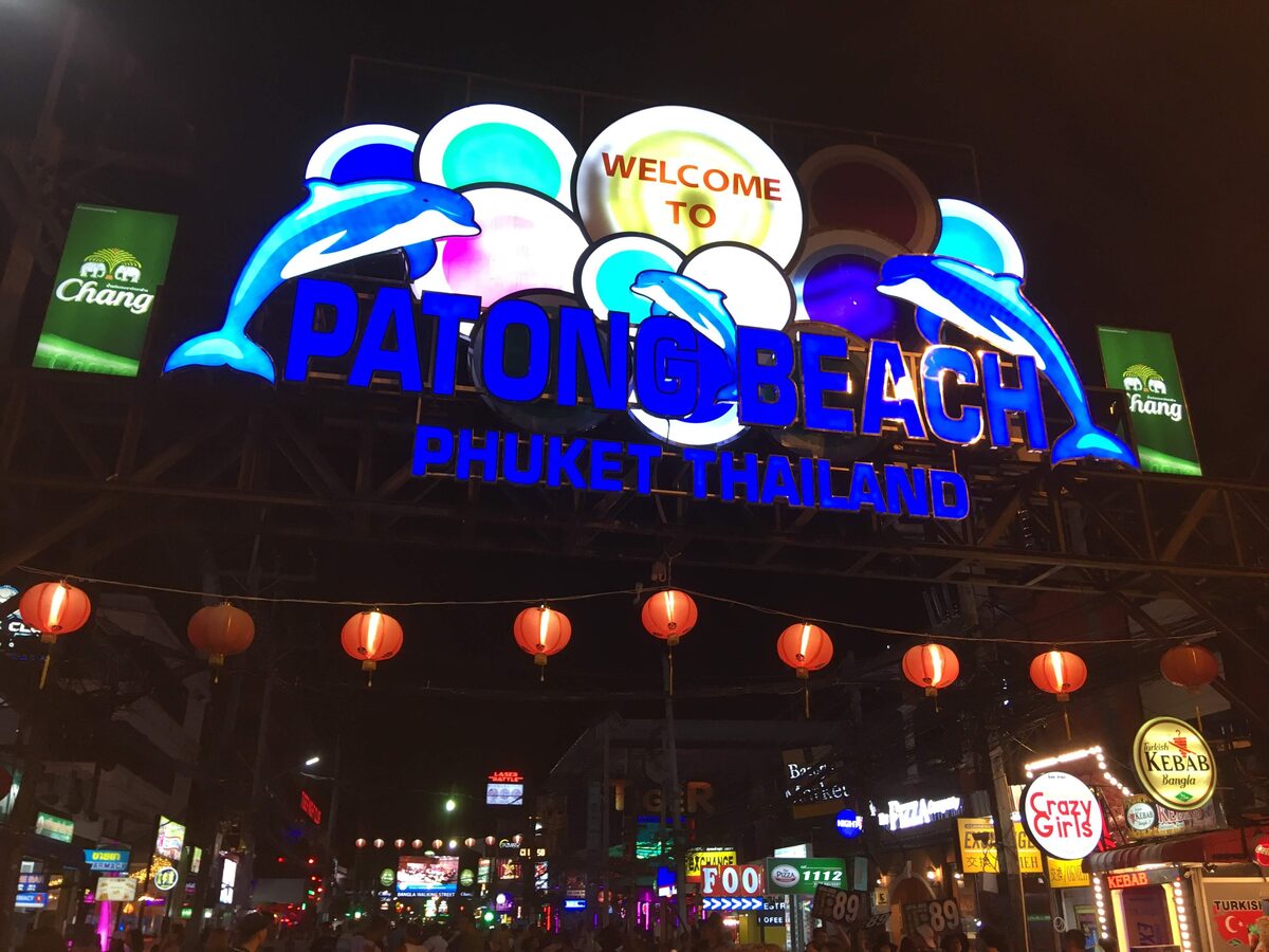 The lit Welcome to Patong Beach sign with dolphins hangs over red paper lanterns at the entrance of Bangla Road at night