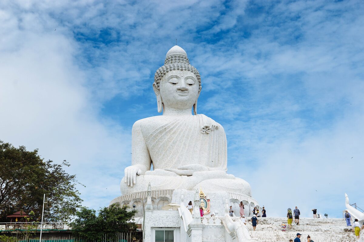 The white seated Big Buddha on its hilltop under a blue sky, with visitors on the steps below