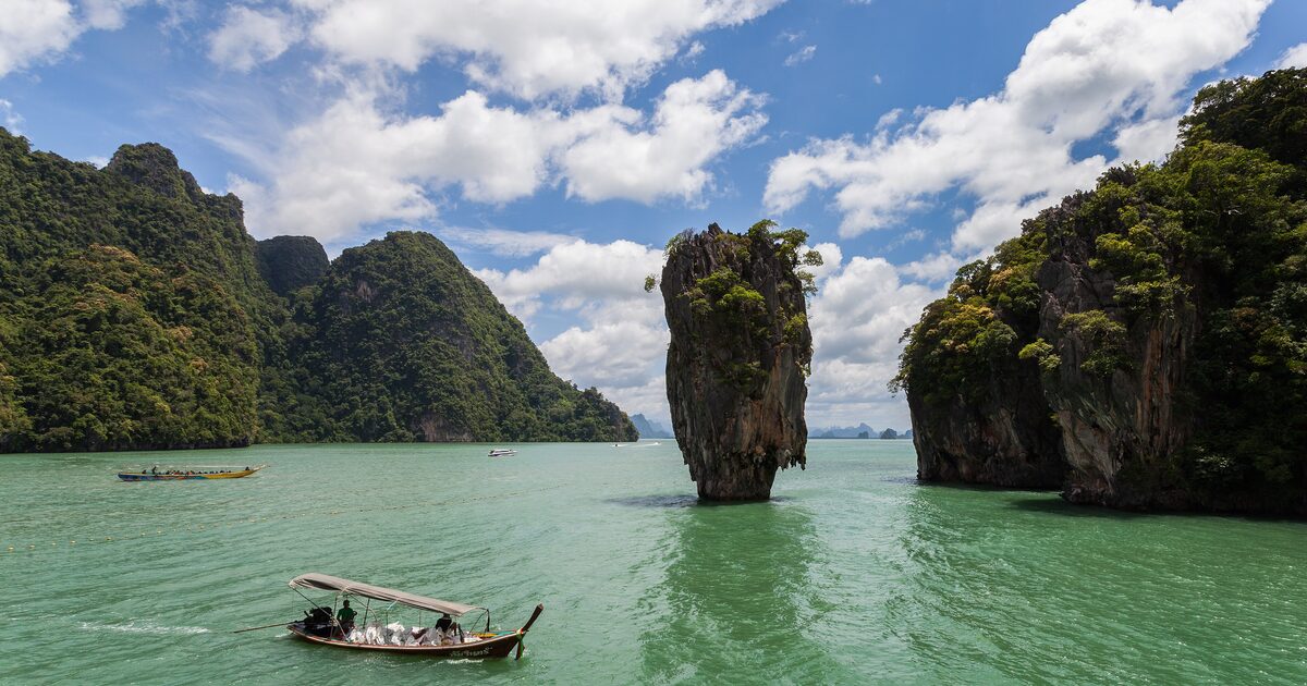 Limestone karsts rise from green water in Phang Nga Bay, with a longtail boat and the pillar of James Bond Island in the middle