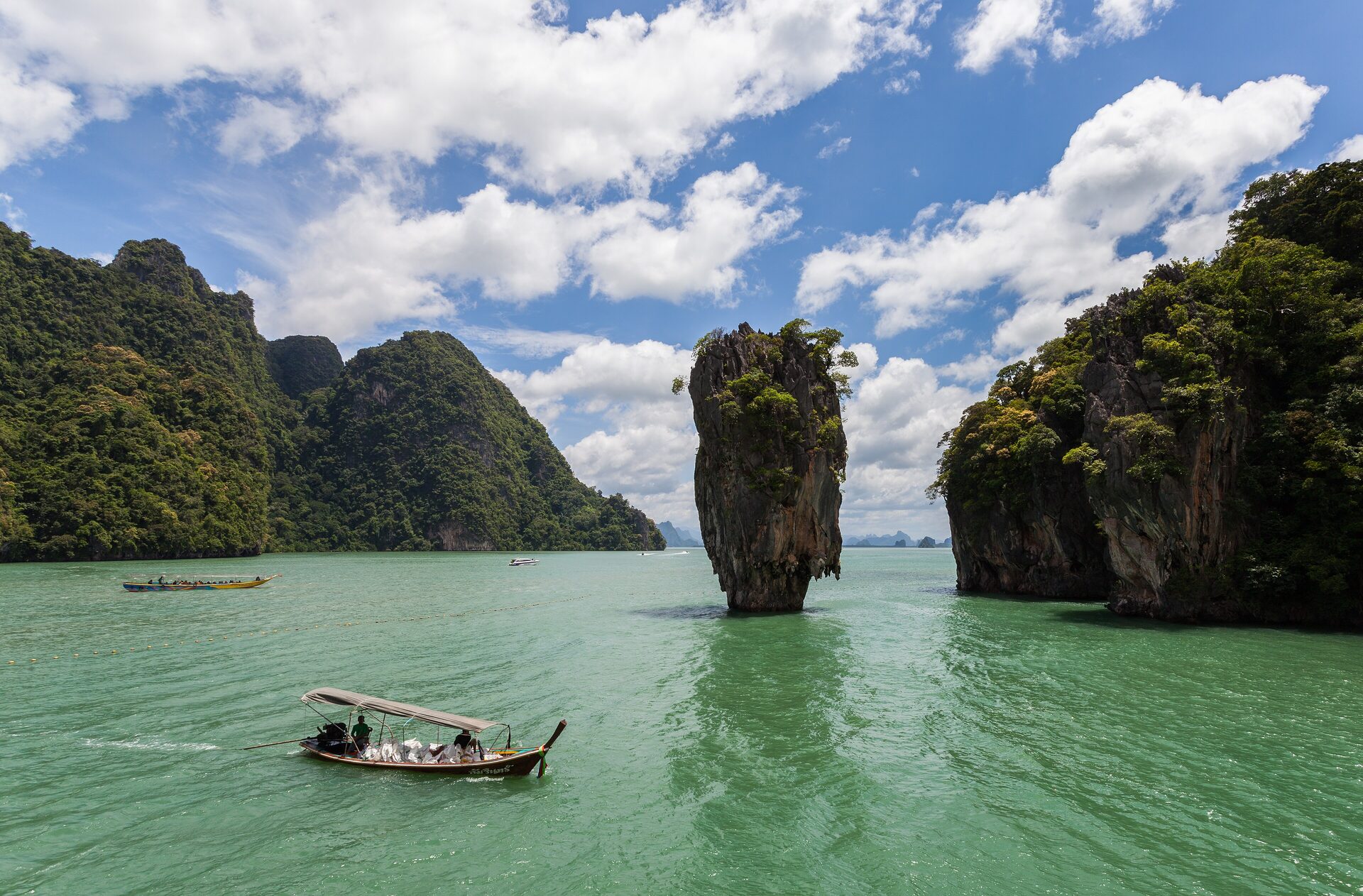 Limestone karsts rise from green water in Phang Nga Bay, with a longtail boat and the pillar of James Bond Island in the middle