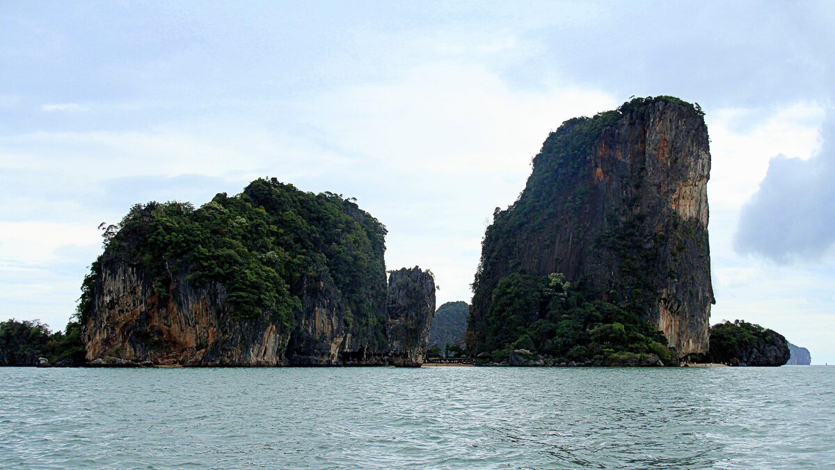 Two limestone pillars rise from calm water at James Bond Island, under a blue sky with clouds