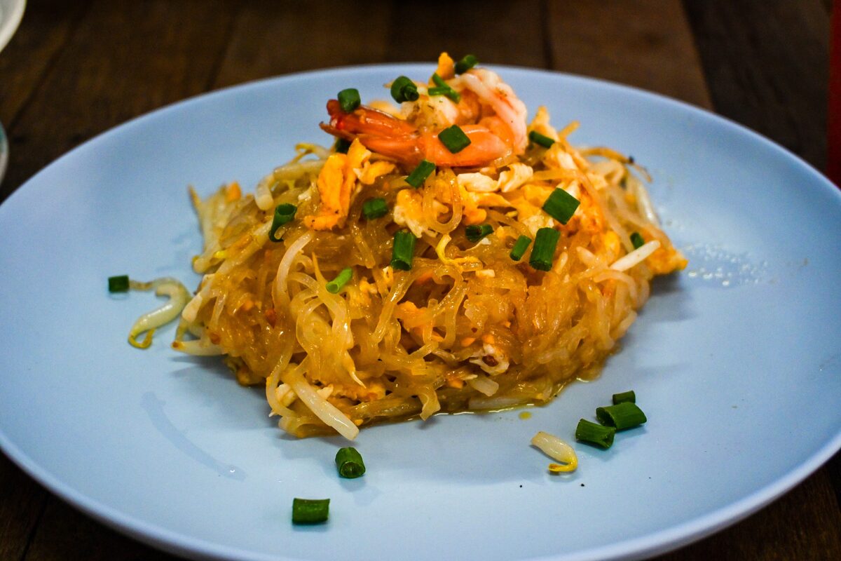 A heap of golden pad thai noodles topped with a shrimp, bean sprouts and chopped scallions on a pale blue plate