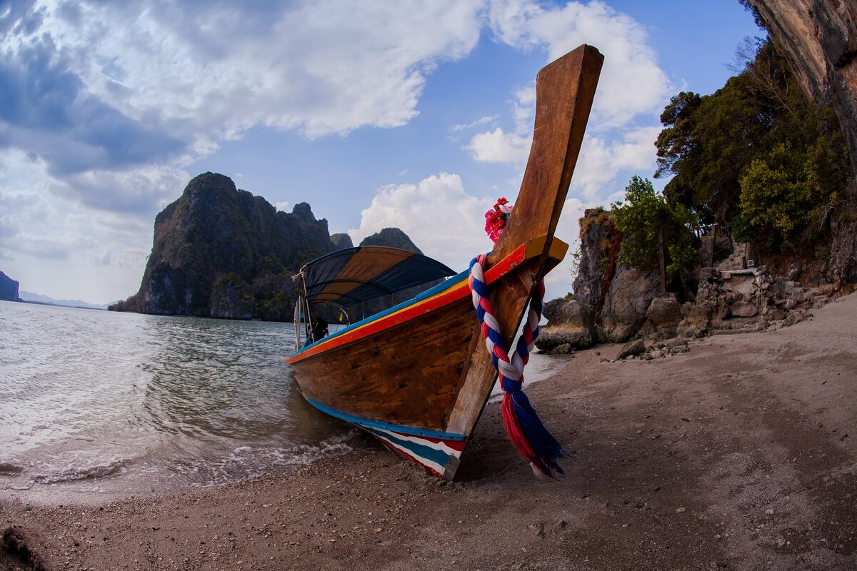 A longtail boat with a colorful ribbon-wrapped bow rests on a small beach in Phang Nga Bay, with limestone karsts behind it