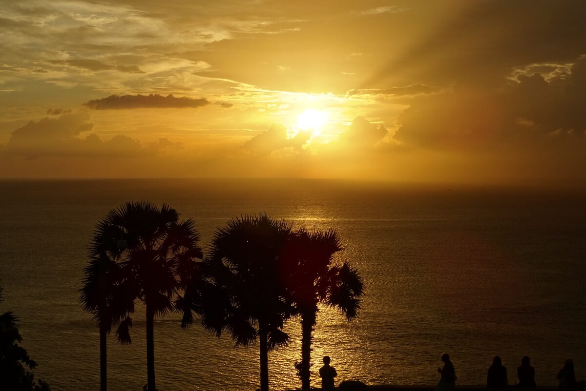 The sun sinks into golden clouds over the Andaman Sea at Promthep Cape, with palm trees and onlookers in silhouette