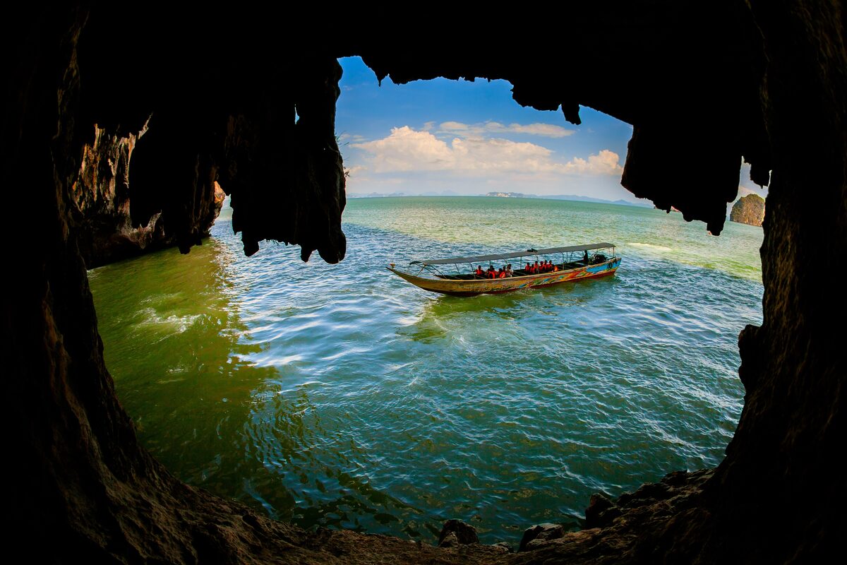 A longtail boat floats in a sea cave, with the open bay and green water framed by dark rock