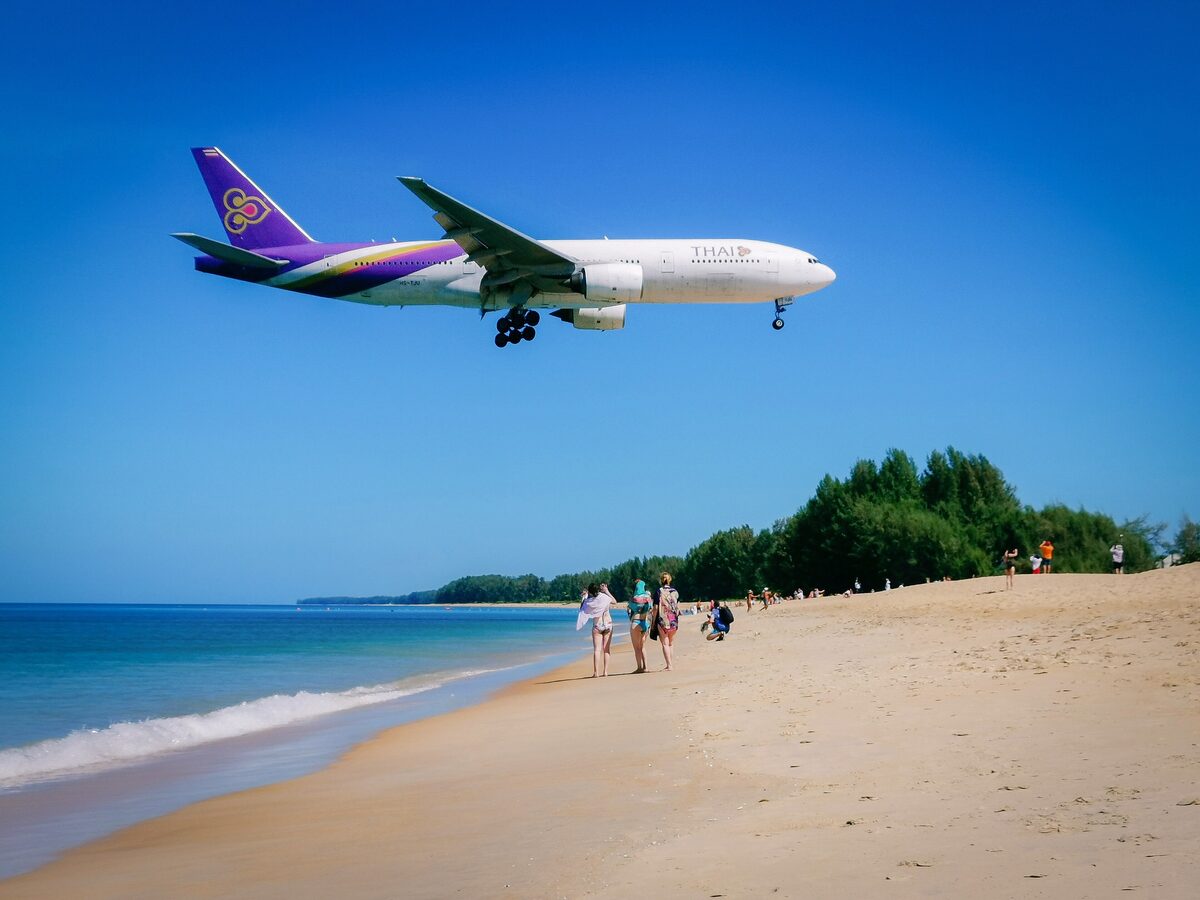 A jet comes in low over Mai Khao beach, with people walking on the sand below
