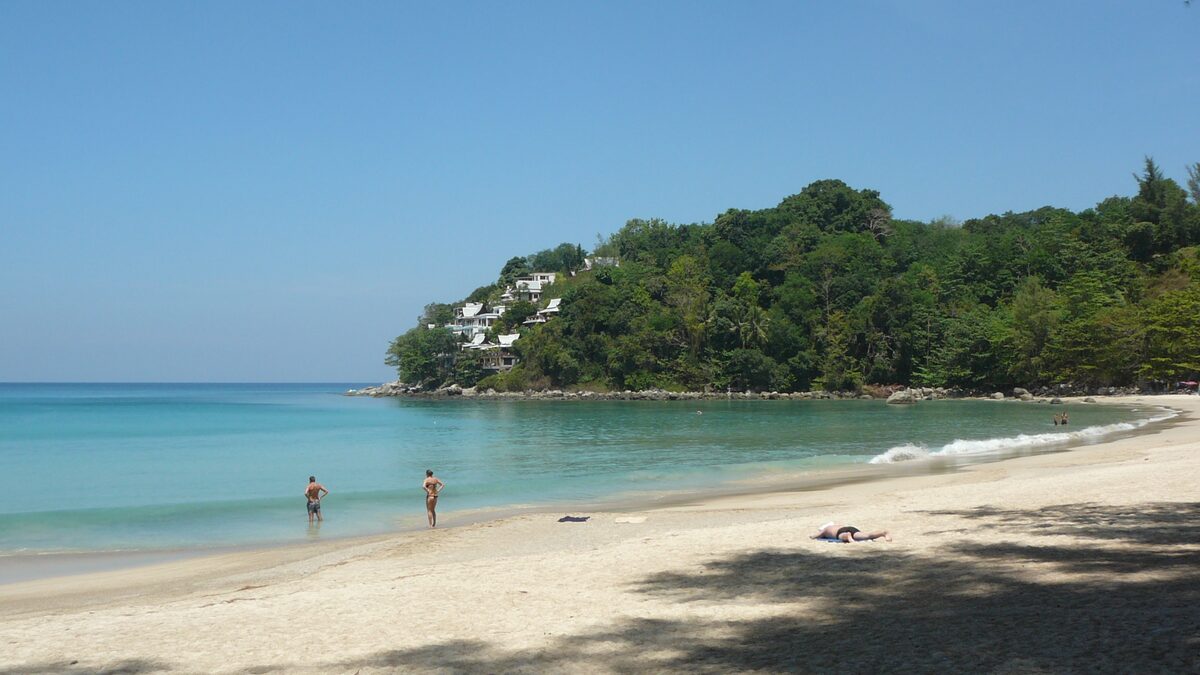 Pale sand and clear water at Kamala Beach, with a wooded headland and a white hillside villa in the distance