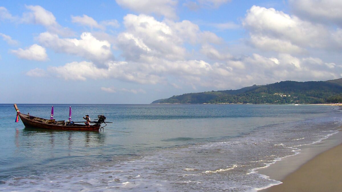 A long-tail boat on the sand at Karon Beach, with a wide bay, clouds and a green headland behind