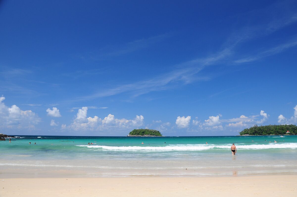 Turquoise water and a white surf line wash onto the pale sand of Kata Beach, with Ko Pu and a second island under a blue sky
