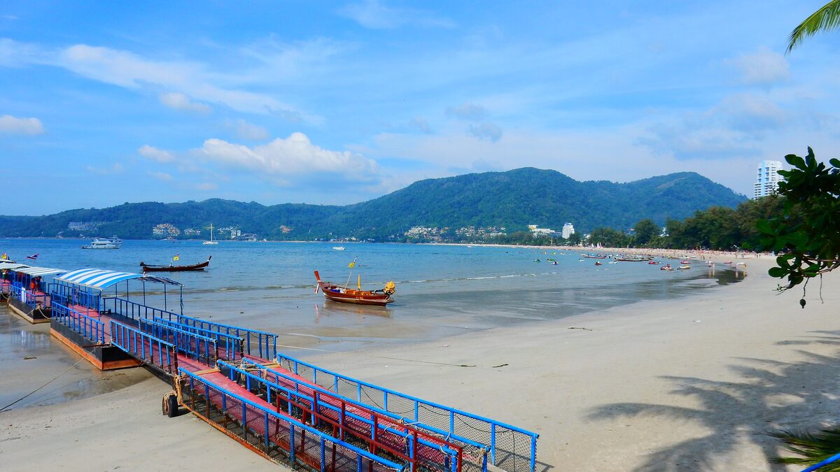 A blue jetty and long-tail boats on the sand at Patong Beach, with hills across the bay