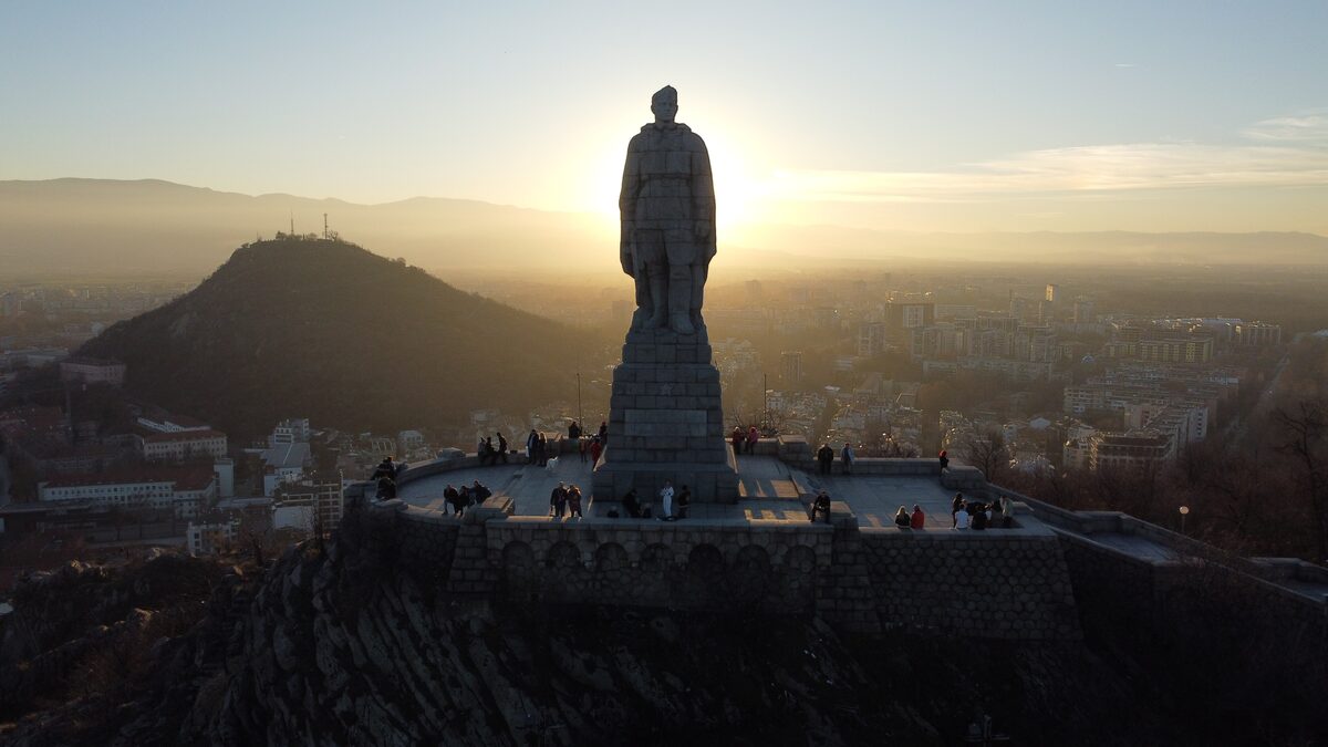 The Alyosha monument on Bunardzhik hill silhouetted against the setting sun, people on the terrace around its base and Mladezhki hill and Plovdiv behind.