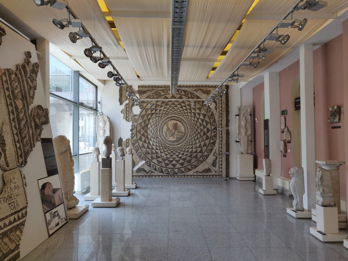 The Roman hall of the Regional Archaeological Museum in Plovdiv, with marble statues and reliefs on plinths and a large geometric floor mosaic mounted on the end wall.