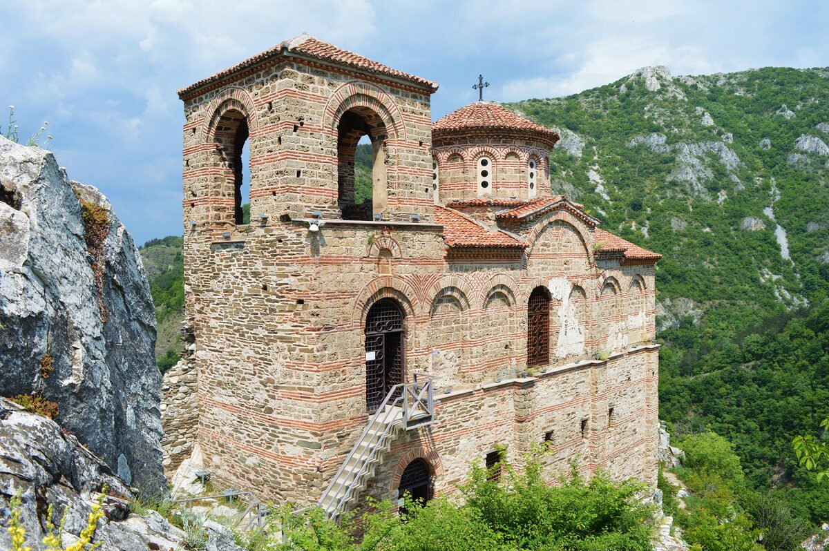 The Church of the Holy Mother of God at Asen's Fortress, a banded stone-and-brick medieval church with a bell tower, on a rocky crag above the green Rhodope slopes.