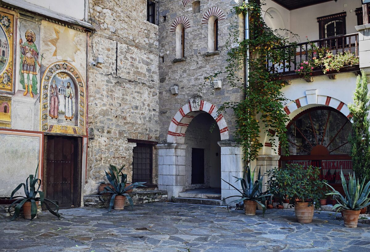 A stone-paved courtyard of Bachkovo Monastery with frescoed saints on the wall, red-and-white striped arches, potted agaves and a flower-hung wooden balcony.