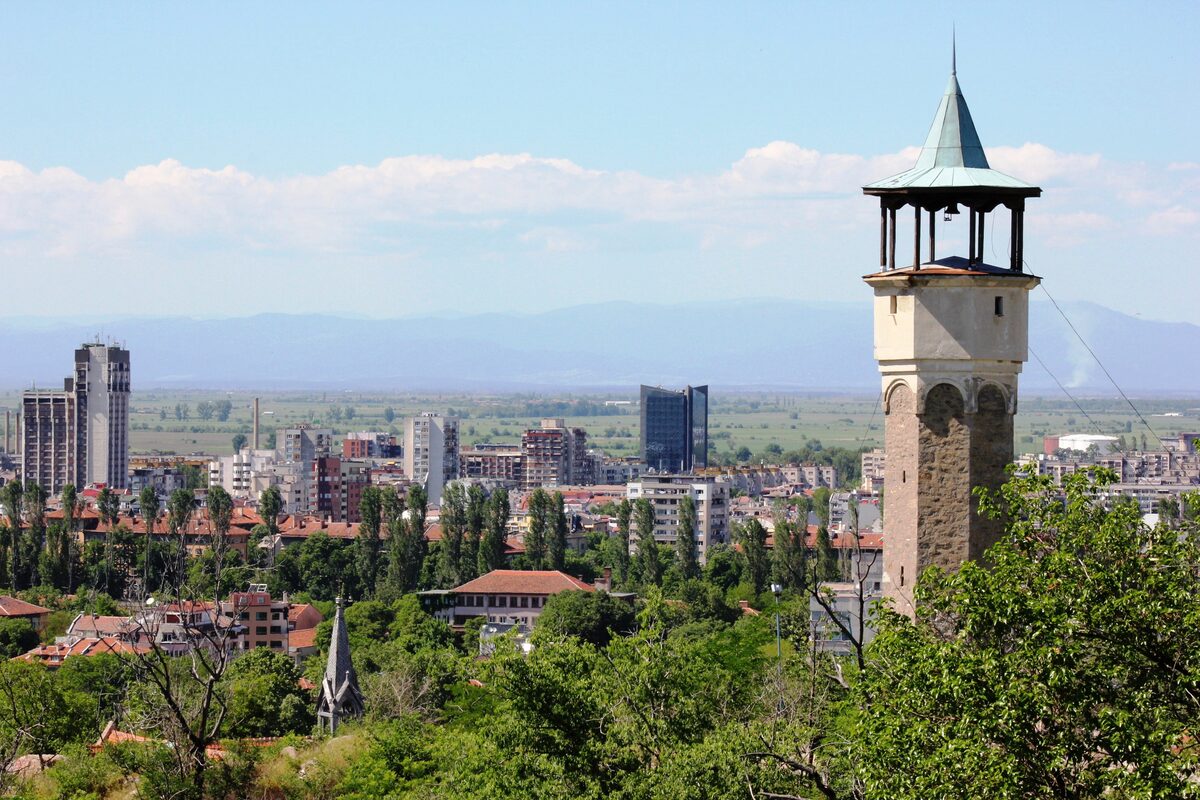 The Ottoman clock tower on Sahat Tepe with its green pointed roof, rising above the trees with Plovdiv and the Thracian plain stretching to distant mountains behind.