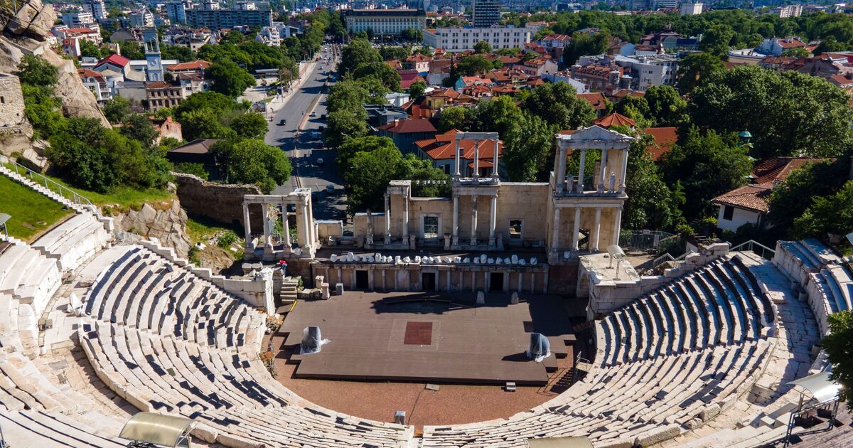 Aerial view of the Ancient Theatre of Philippopolis with its marble seating and two-storey stage front, the red roofs of Plovdiv and a long boulevard behind it.