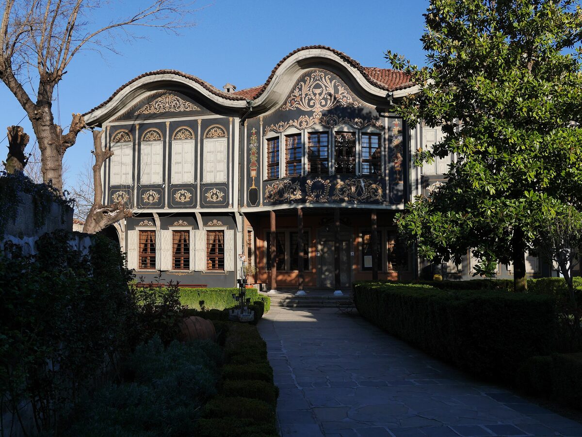 The Kuyumdzhiev House, home of the Regional Ethnographic Museum, with its wavy baroque roofline and dark blue facade painted with gold garlands, seen down a garden path.