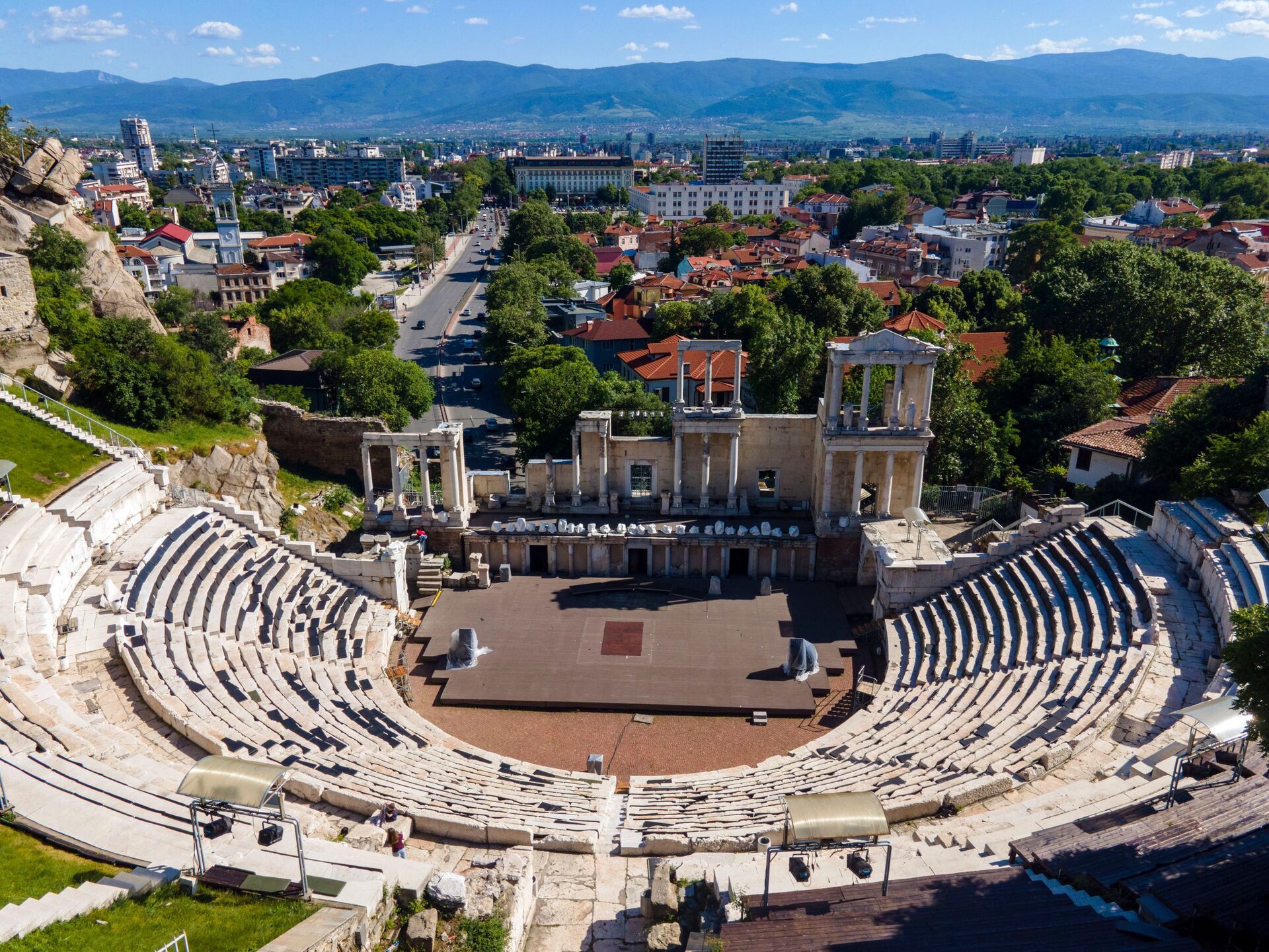 Aerial view of the Ancient Theatre of Philippopolis with its marble seating and two-storey stage front, the red roofs of Plovdiv and a long boulevard behind it.