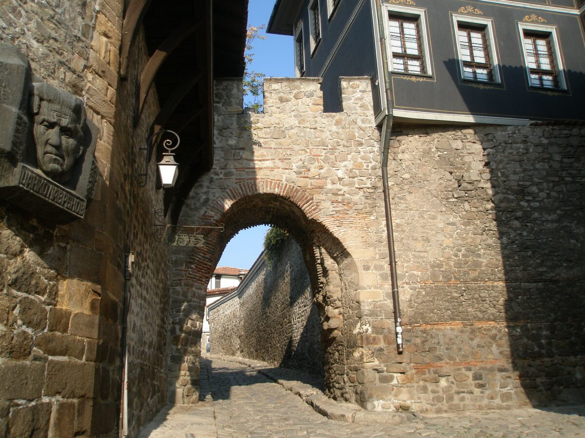 The stone arch of Hisar Kapiya over a cobbled lane in Old Plovdiv, a dark blue Revival house built on top of the old fortress wall.
