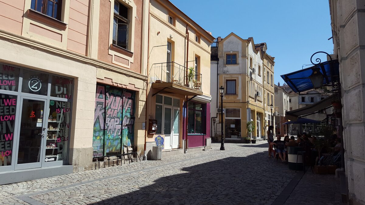 Cobbled Zlatarska street in the Kapana district, with a graffiti-covered shop shutter, small boutiques and people at cafe tables in the shade.