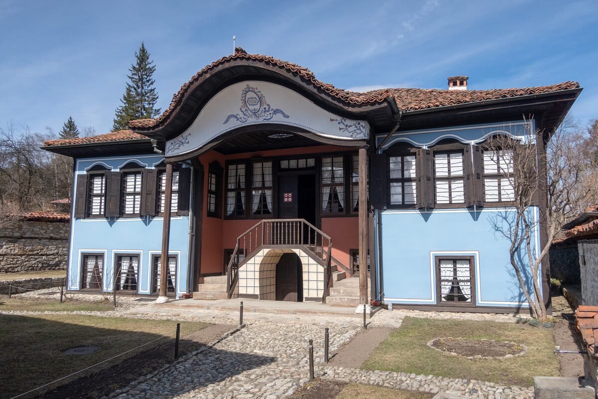 Lyutov House in Koprivshtitsa, a sky-blue Revival-era house with a curved, painted gable over the porch, dark wooden shutters and a cobbled garden path.