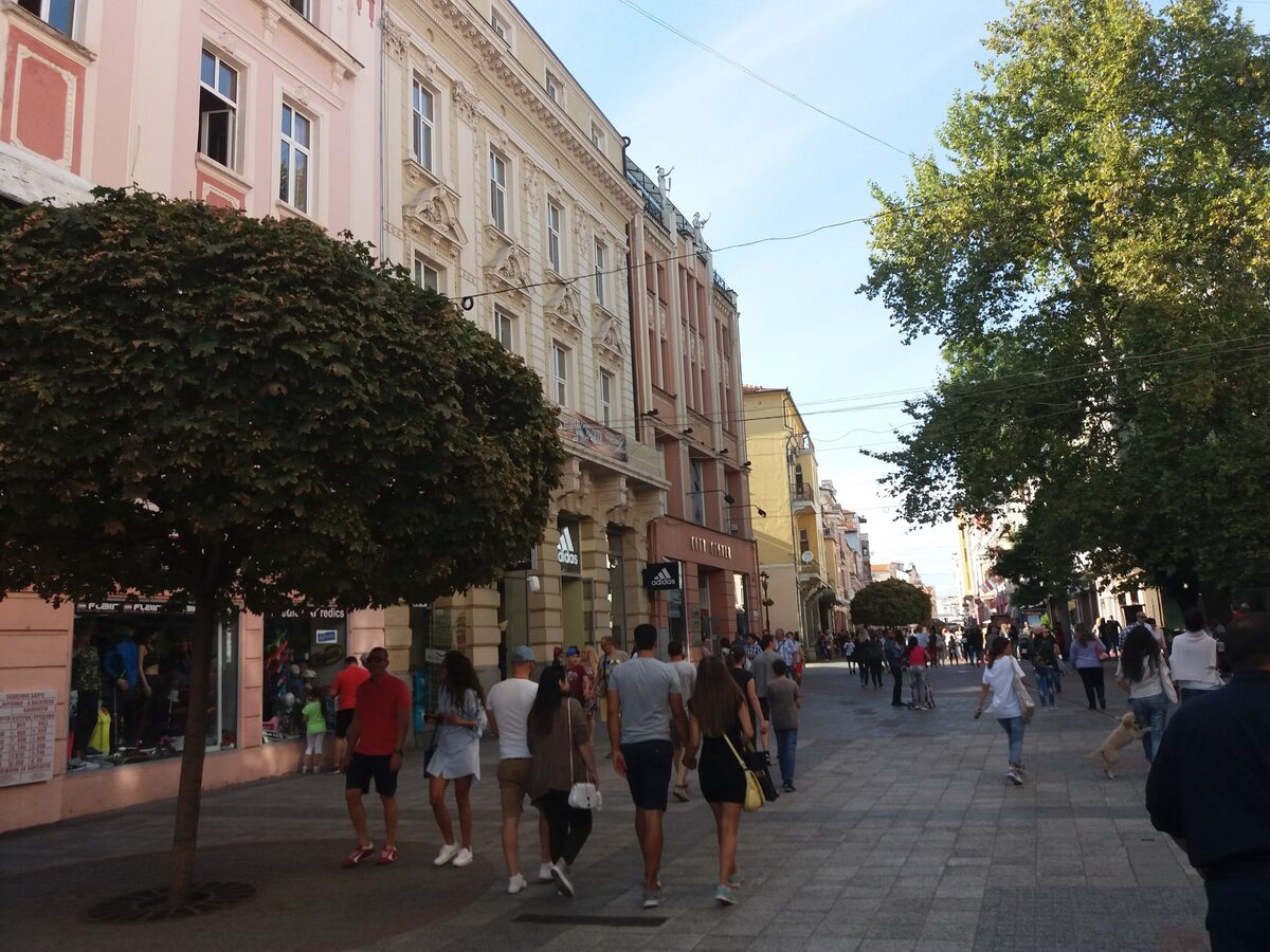 Strollers on the pedestrian Knyaz Alexander I street, lined with trees and pastel late 19th-century facades.