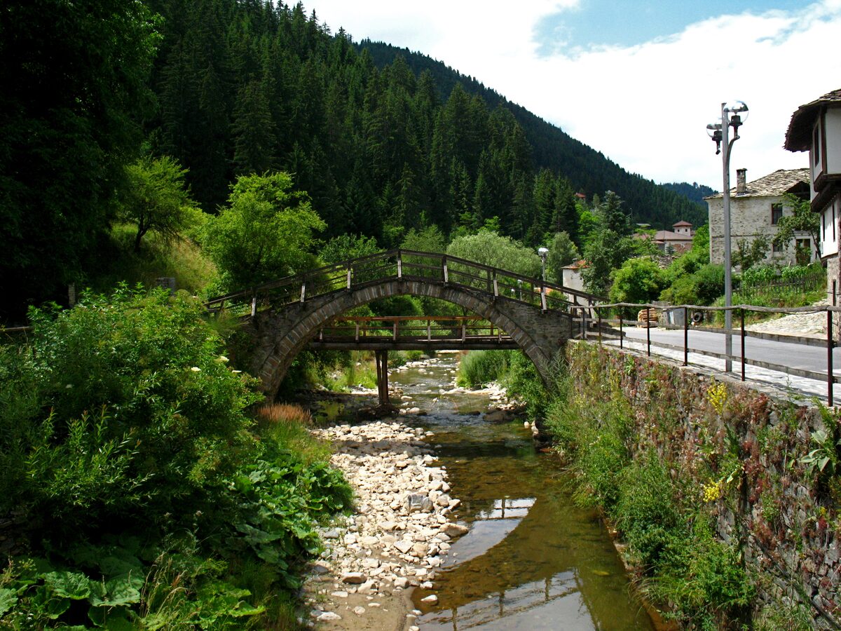 An arched stone footbridge over the shallow stream in Shiroka Laka, with stone-roofed houses on the bank and spruce-covered Rhodope slopes behind.