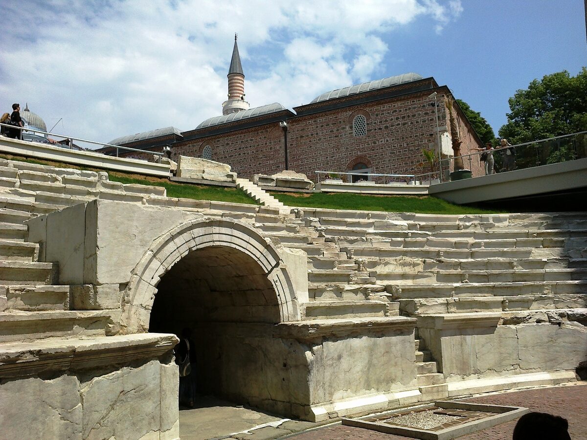 The curved end of the Roman Stadium of Philippopolis with its arched entrance and marble seats, the brick walls and minaret of the Dzhumaya Mosque rising above.