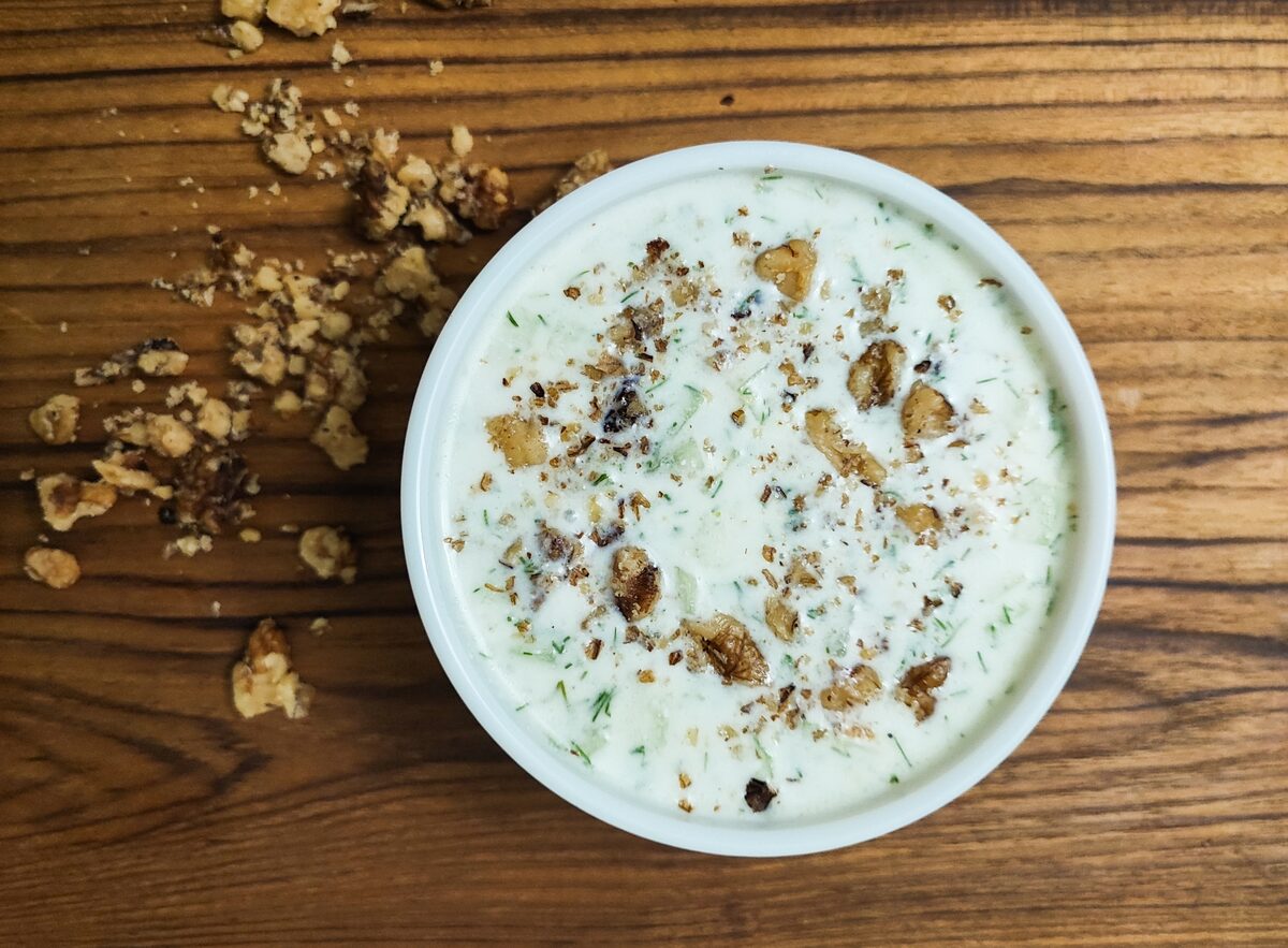 A white bowl of cold tarator with yogurt, cucumber and dill, sprinkled with crushed walnuts, on a wooden table.