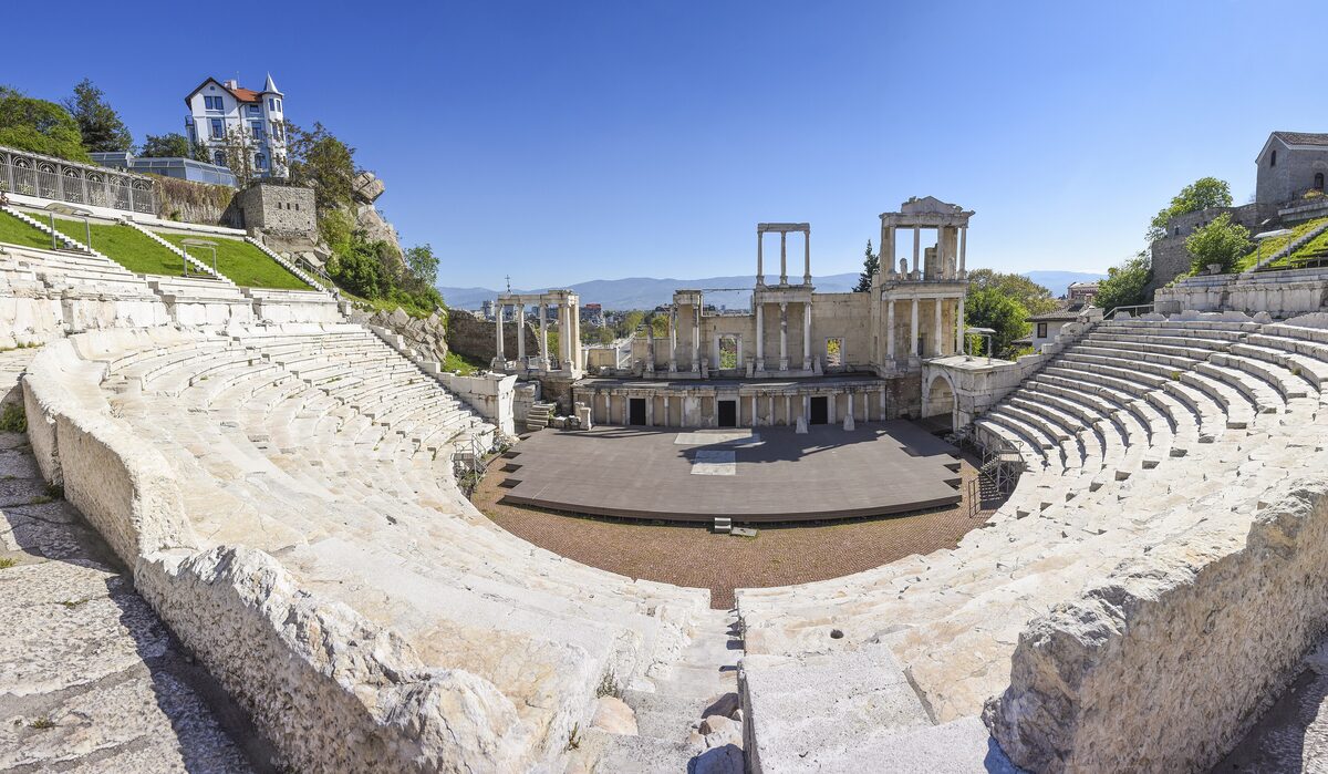 Wide view from the upper rows of the Ancient Theatre of Philippopolis, the white marble tiers curving down to the columned stage under a clear blue sky.