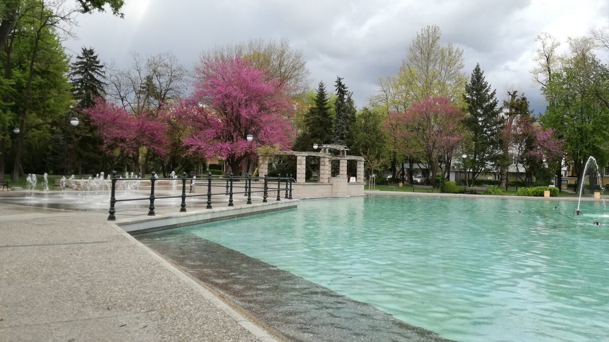 The turquoise pool of the Singing Fountains in Tsar Simeon Garden in spring, with pink-flowering Judas trees, a stone pergola and small fountain jets.