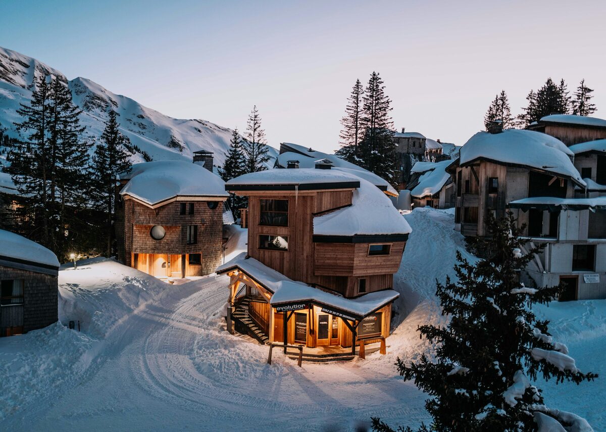 Wooden Avoriaz chalets lit up at dusk on a snowy street, with a peak behind