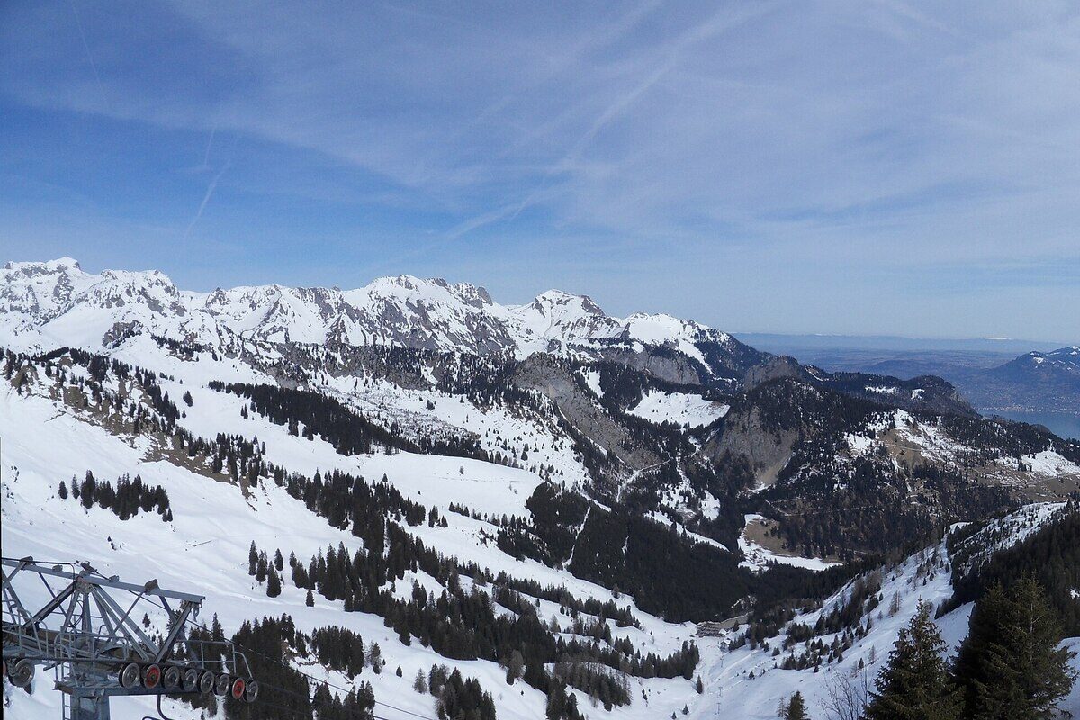 Snowy ridges and forested slopes seen past a lift station above Châtel, with Lake Geneva faint on the horizon