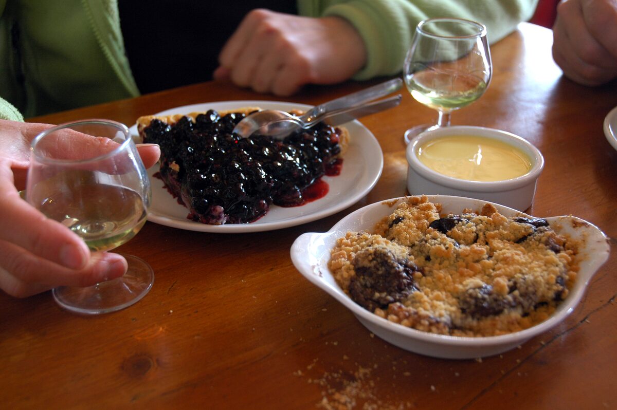 Small glasses of pale liqueur on a wooden table beside a blueberry tart and a fruit crumble
