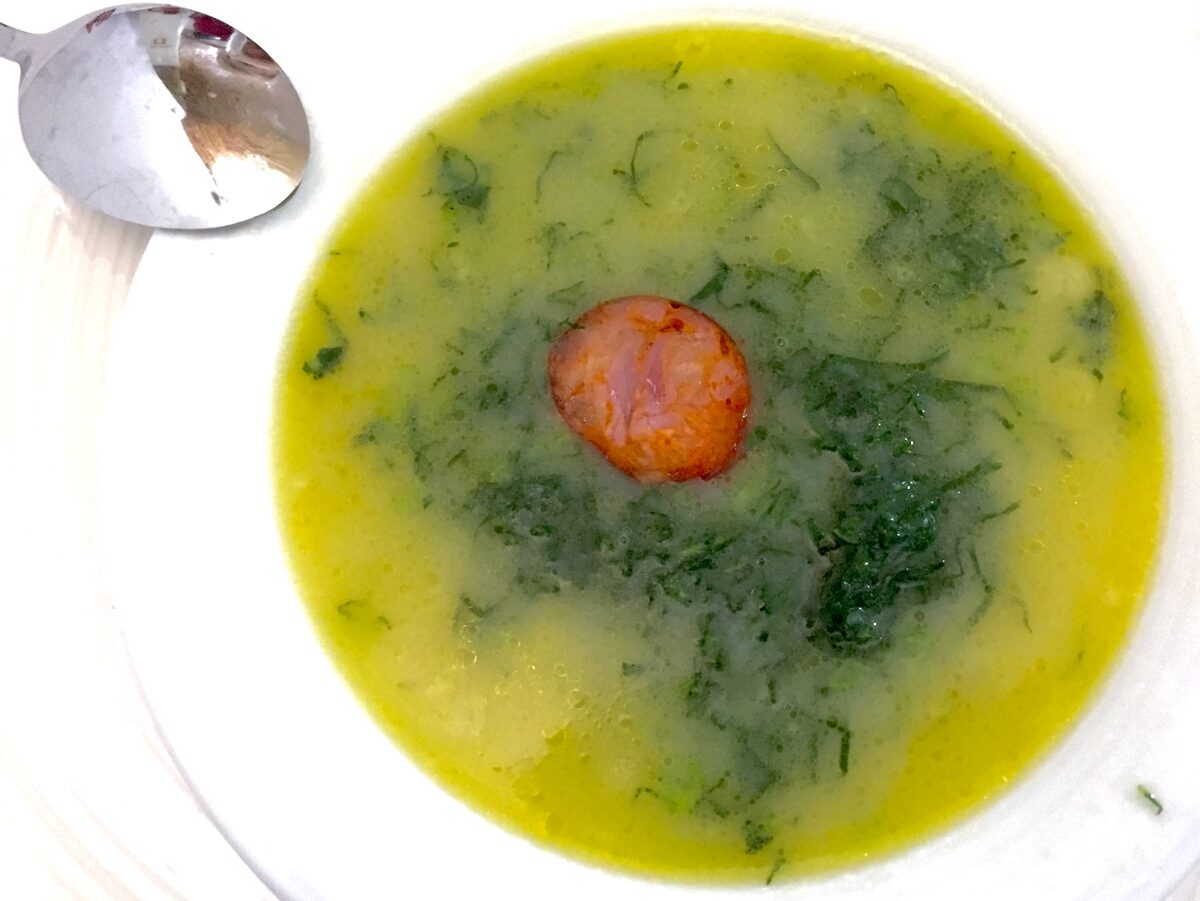 A white bowl of caldo verde, yellow-green potato soup with shredded kale and a slice of chorizo