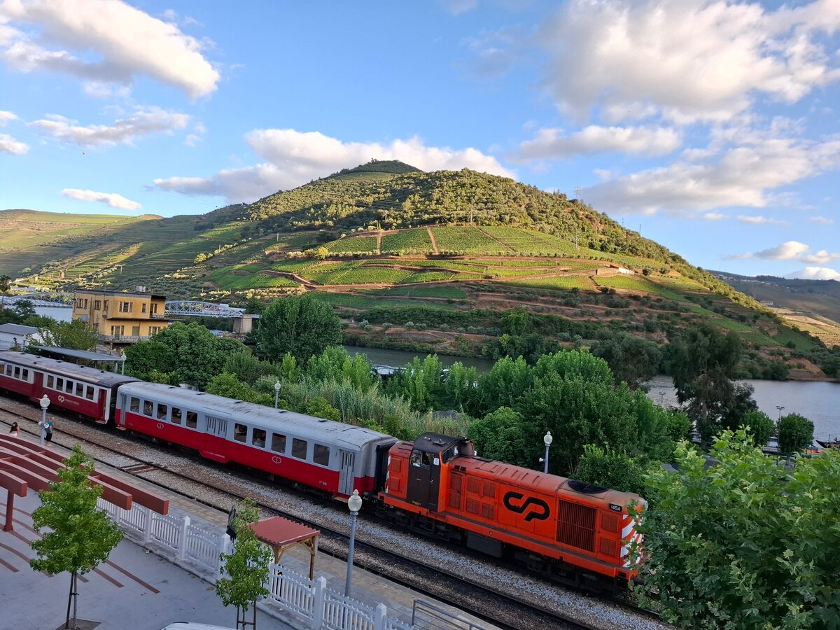An orange CP locomotive pulling a red and grey train along the river at Pinhão, with terraced vineyards on the hill behind