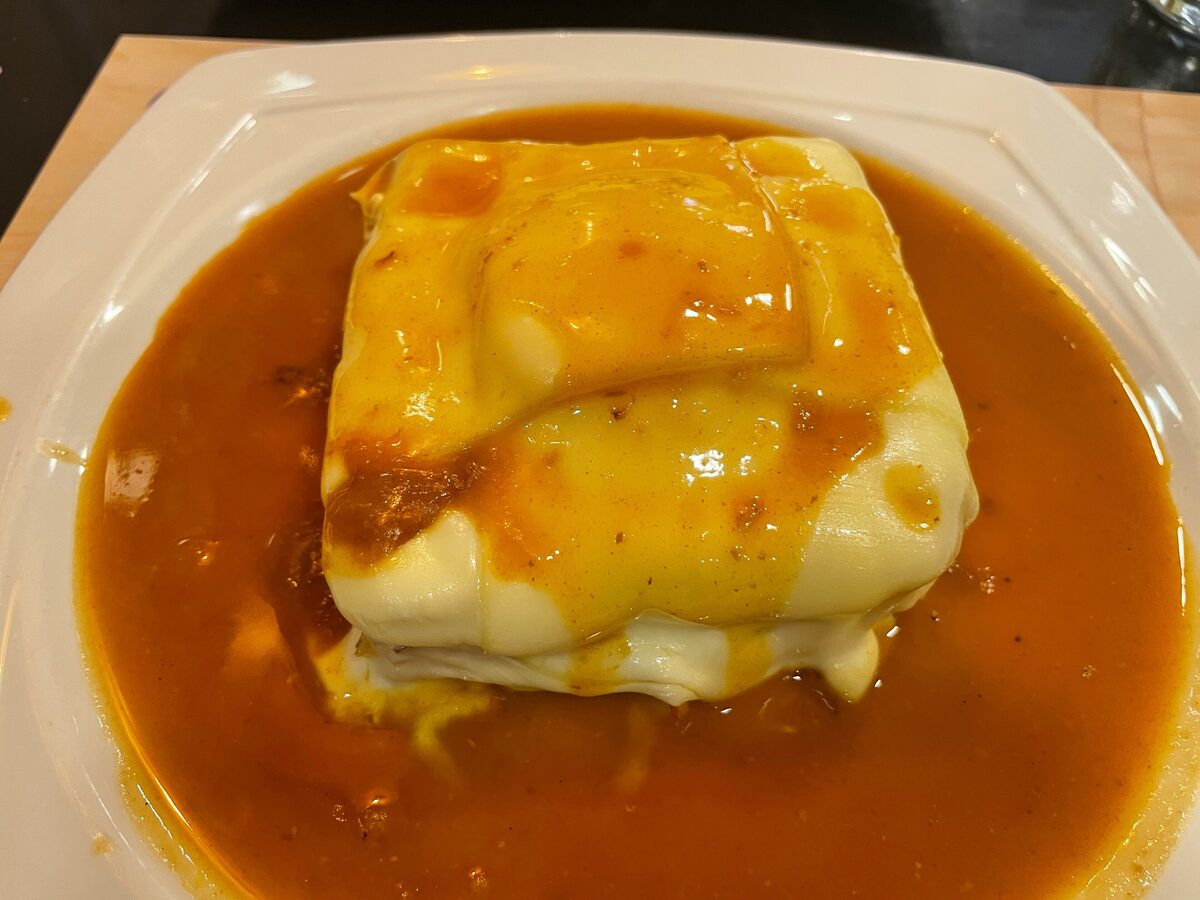A francesinha, a sandwich buried under melted cheese, sitting in a bowl of orange tomato and beer sauce