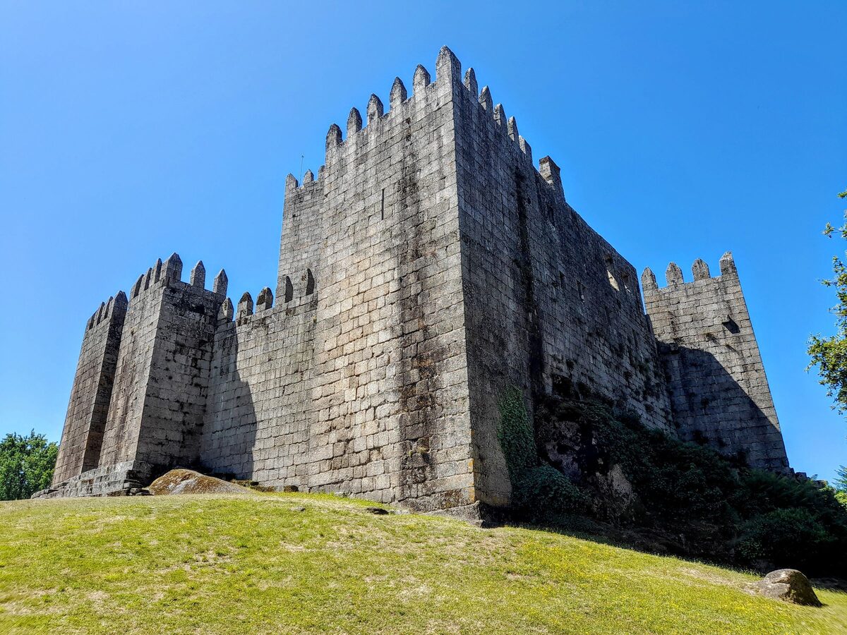 The granite keep and crenellated walls of Guimarães Castle above a green lawn under a blue sky