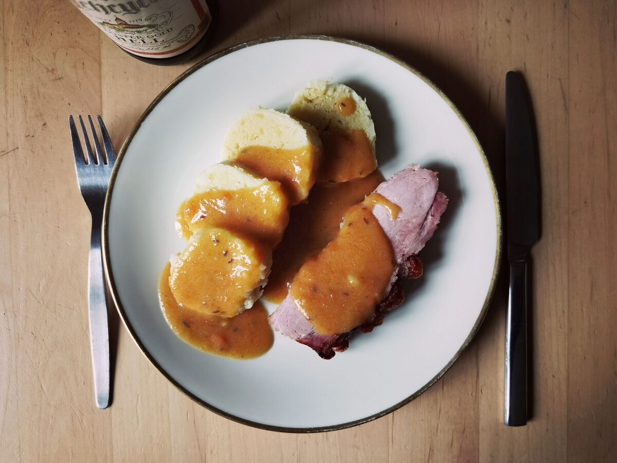 Sliced bread dumplings under gravy next to a slice of roast pork on a white plate