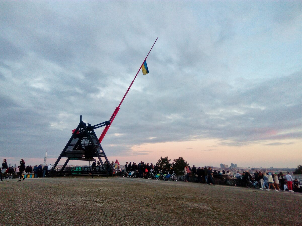 The red pendulum of the Prague Metronome on Letna hill at dusk, with a crowd along the wall