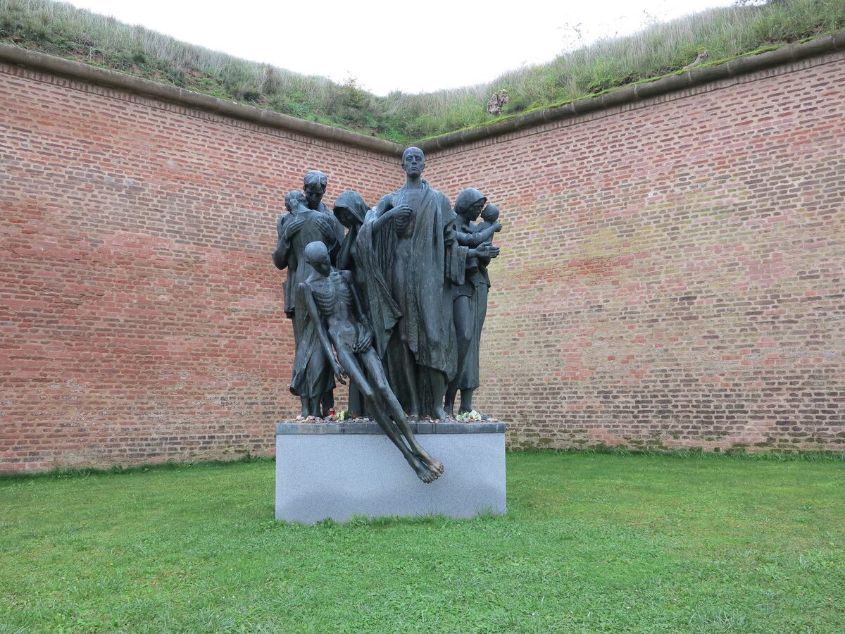 Bronze memorial figures in the Small Fortress at Terezín, set against a brick wall and a grass lawn