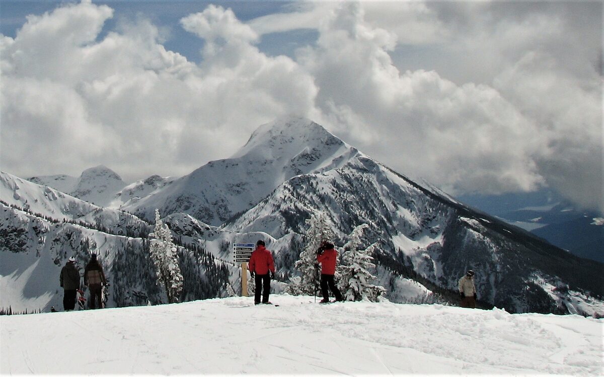Skiers pause at a trail sign facing a pointed snowy peak