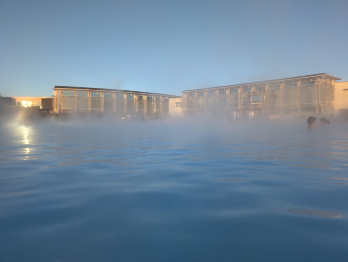 Steam over the blue water of the Blue Lagoon at low light, with the spa buildings behind and two bathers at the right edge