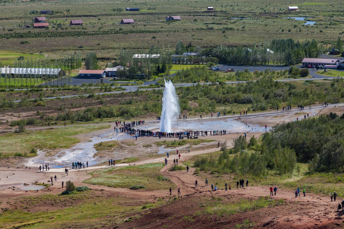 Strokkur erupting in a column of water while a crowd stands around the pool, seen from the hill above the Haukadalur geothermal field