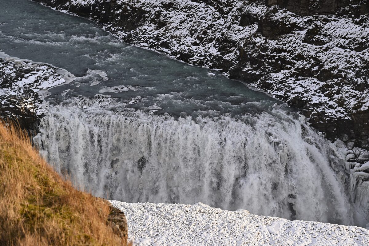 Gullfoss seen from above as the river drops over the edge, with snow and ice on the rocks and a patch of dry grass in the corner