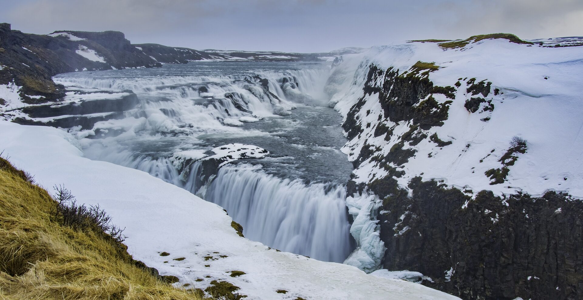 Gullfoss in winter, the river pouring over snow-covered black rock under a pale sky