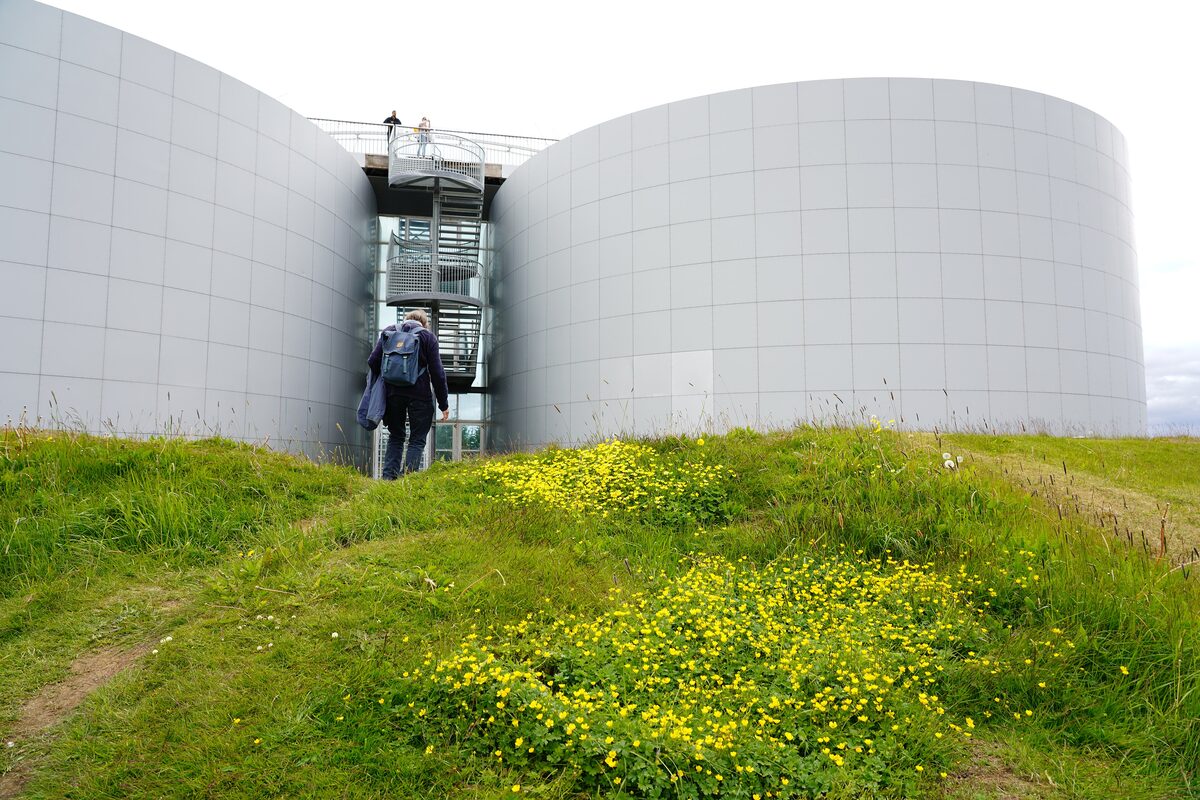A visitor walking up the grass toward Perlan's grey-white tanks, with a metal staircase between them leading to the viewing deck