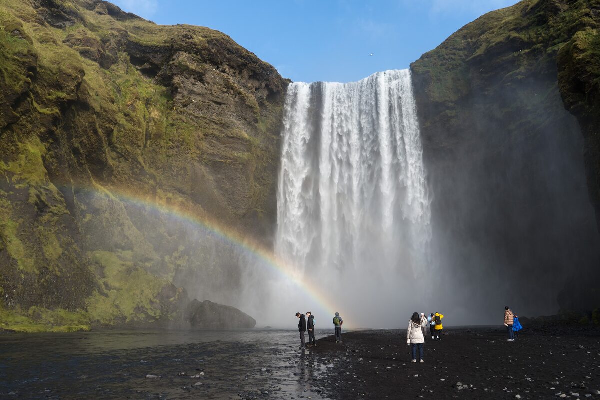 Skógafoss falling over a mossy cliff with a rainbow in the spray, and a few visitors on the black sand below